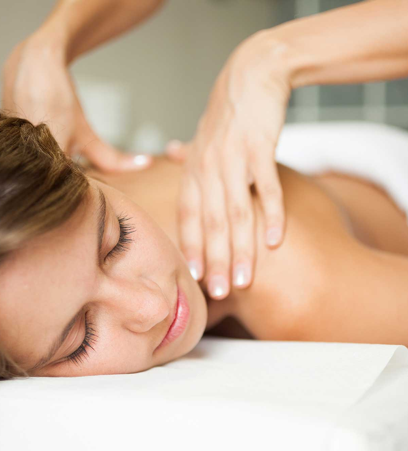 A woman lies face down with eyes closed, relaxing while receiving a shoulder massage from a pair of hands in a softly lit spa setting.