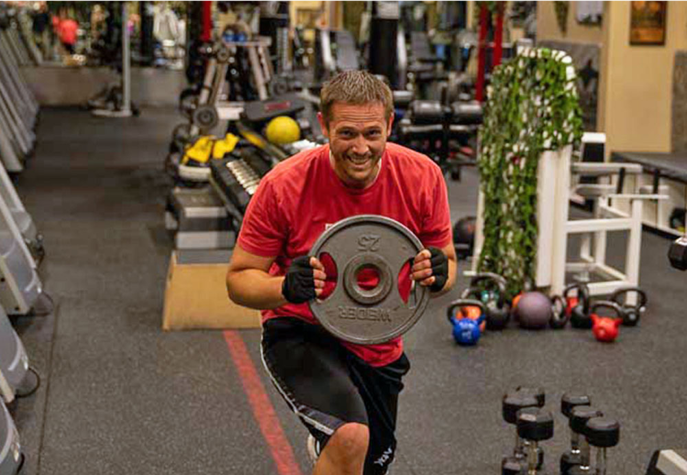 A smiling man in a red shirt and black shorts holds a large weight plate at a gym, surrounded by various exercise equipment including dumbbells, kettlebells, and treadmills.