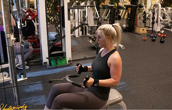 A woman with blonde hair in a ponytail performs a seated cable row exercise in a gym filled with various workout equipment. She wears a dark tank top, black leggings, and weightlifting gloves.