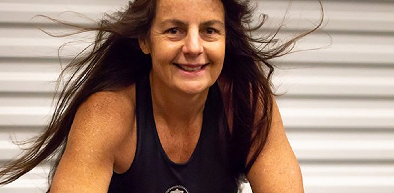 A woman with long brown hair and a sleeveless black top smiles while looking forward, with a blurred metal background behind her.
