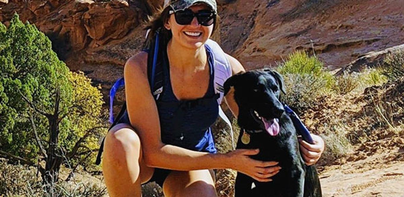 A woman wearing sunglasses and a cap kneels outdoors on a rocky trail, smiling and holding a black dog. The landscape is sunny with shrubs and red rock formations in the background.