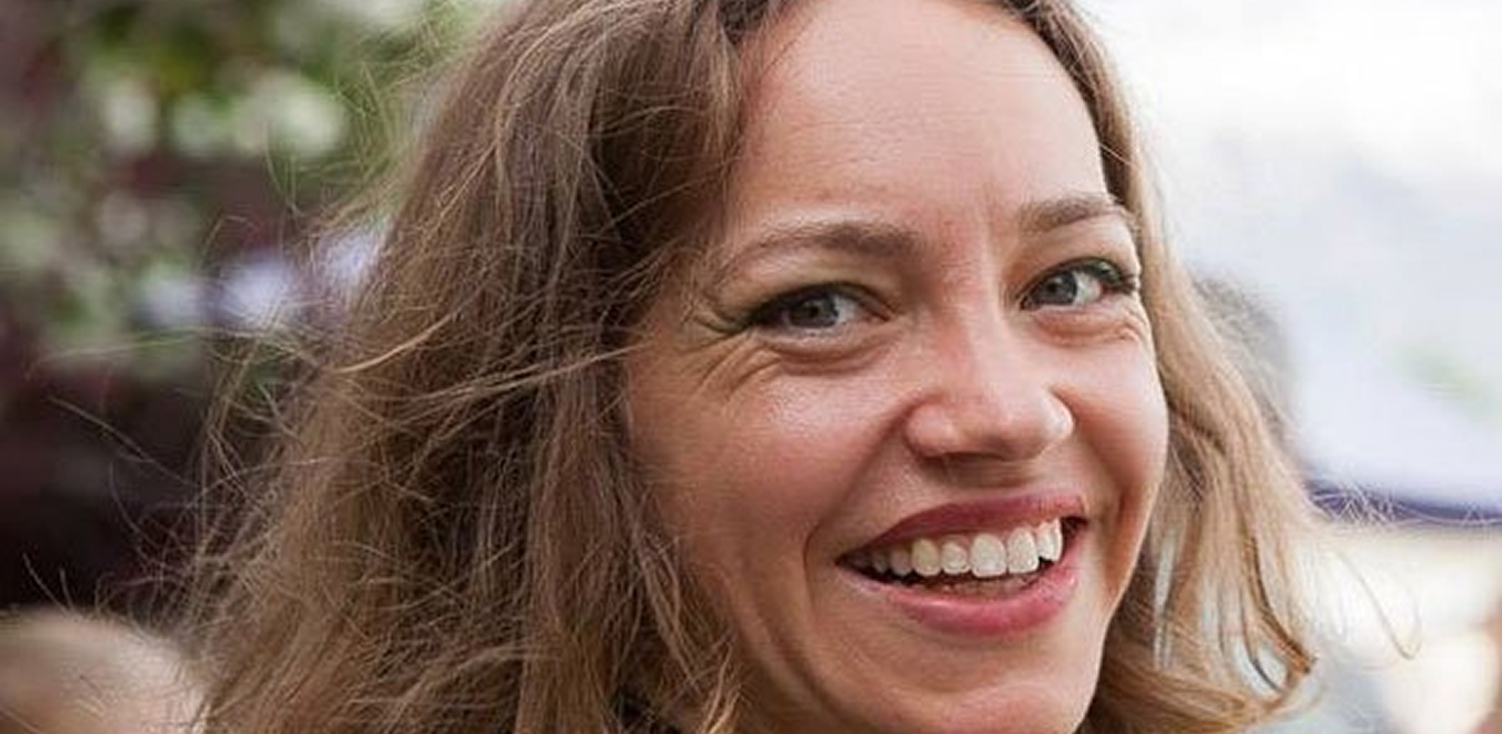 A woman with long brown hair smiles warmly outdoors, with trees and a bright sky blurred in the background.