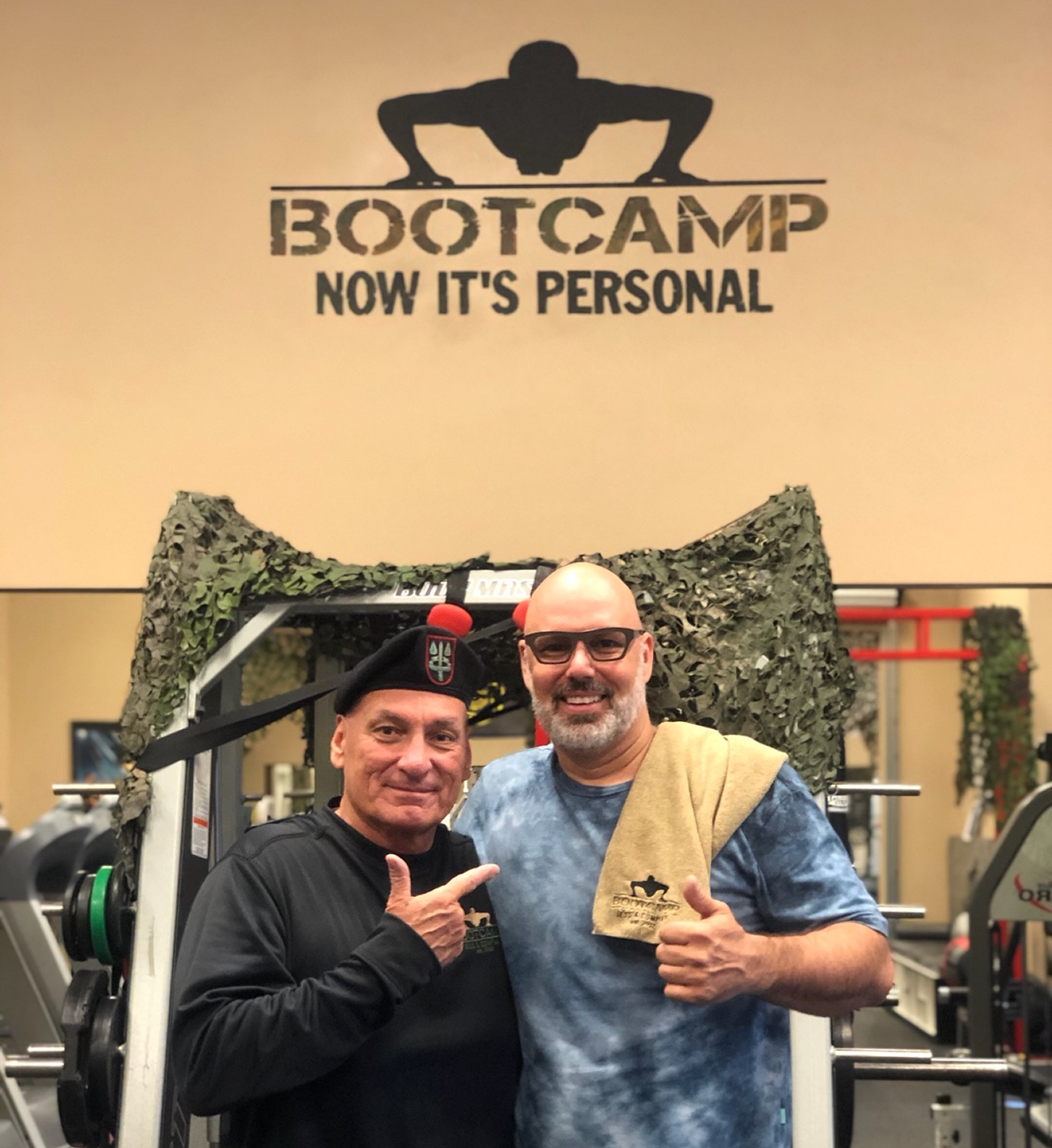 Two smiling men pose at a gym, one wearing a beret and black shirt, the other with glasses and a towel over his shoulder. Behind them is a sign reading "BOOTCAMP NOW IT'S PERSONAL" with a push-up figure logo.
