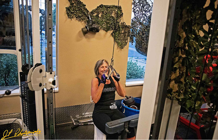 A woman with gray hair smiles while using a cable machine in a gym. She wears a black tank top and blue gloves. Camouflage netting hangs on the walls and equipment surrounds her in the workout area.