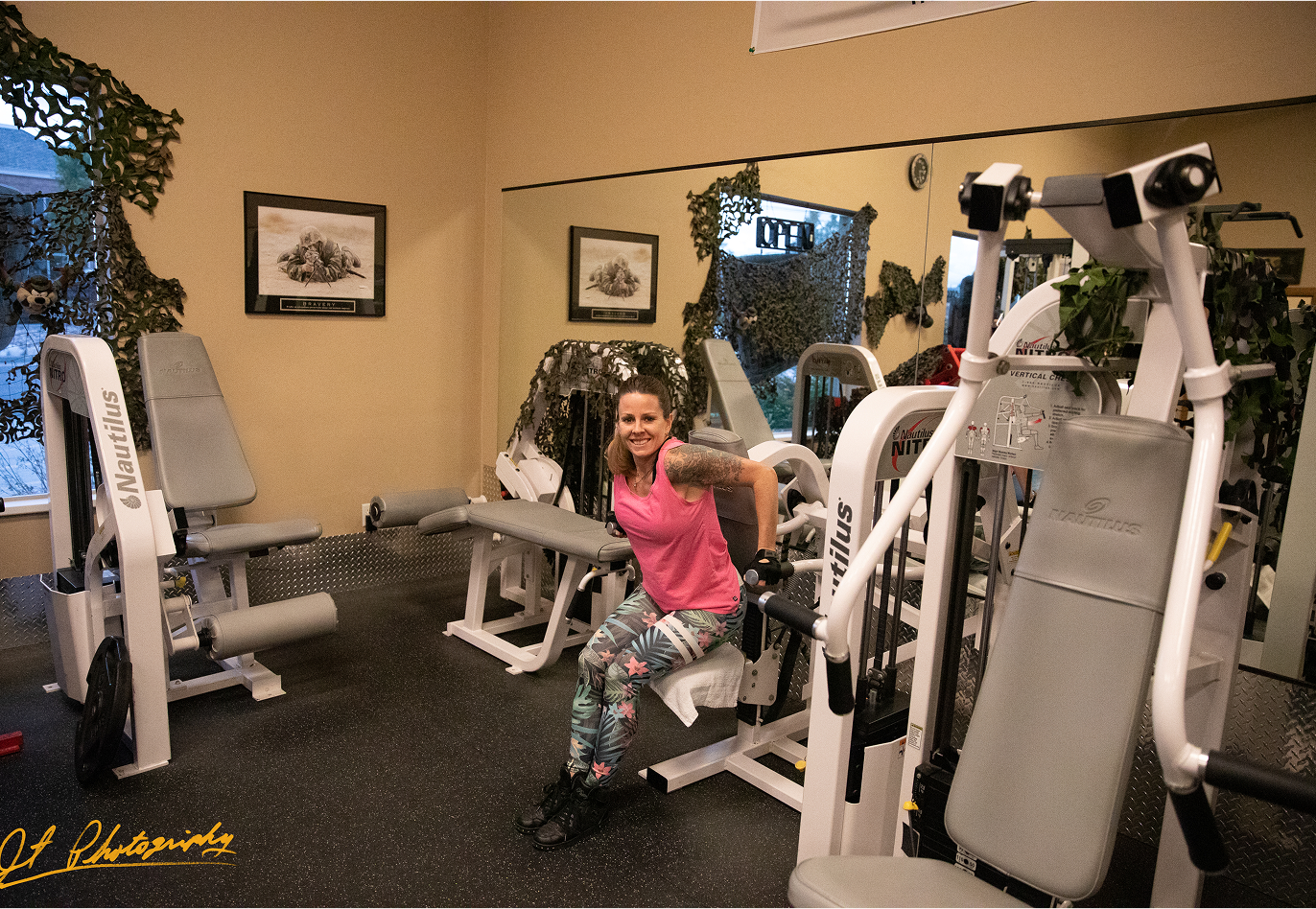 A woman in workout clothes smiles while sitting on a machine in a gym filled with exercise equipment. The room has mirrors, framed photos, and camouflage decorations on the windows.