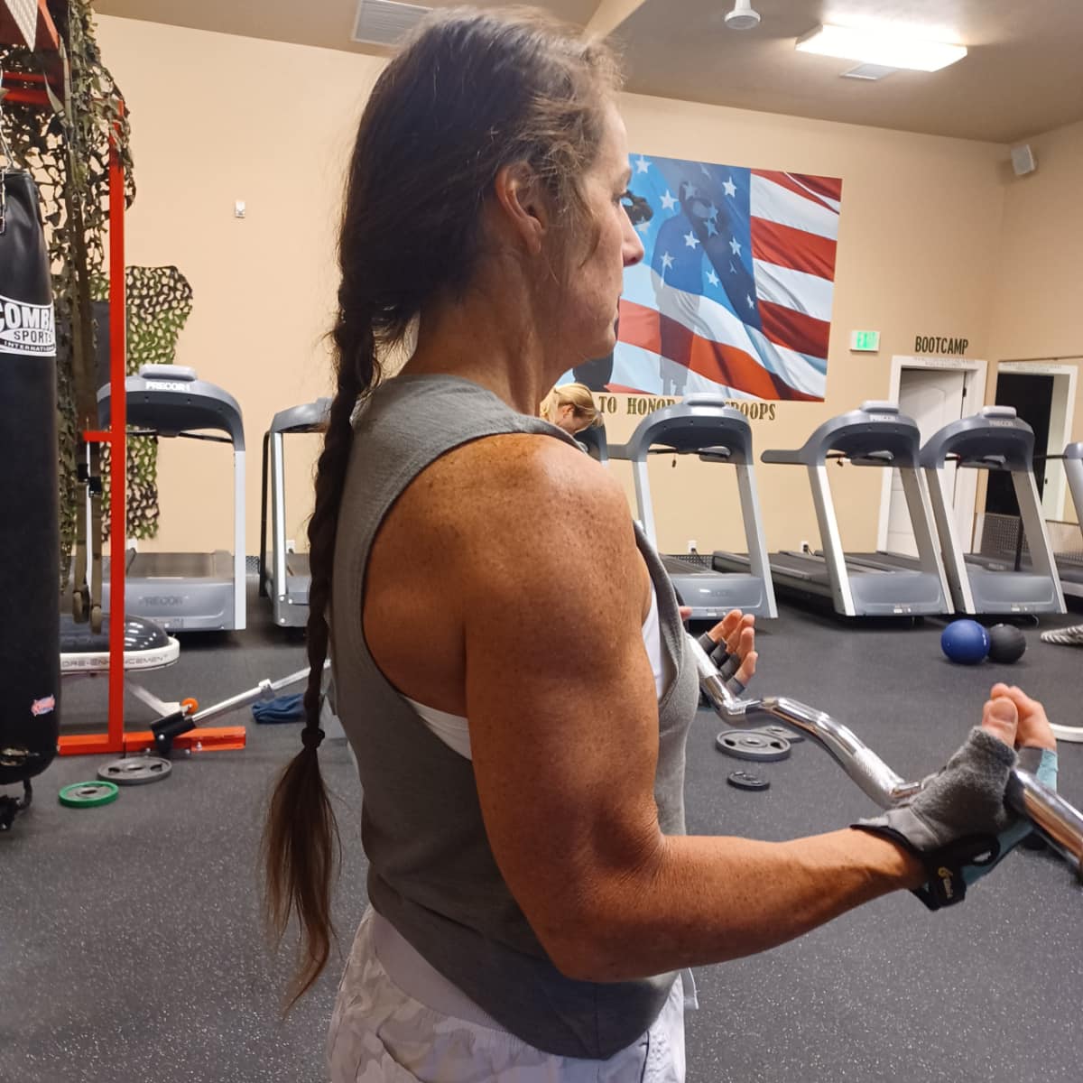 A woman with a long braid works out with a barbell in a gym. She is wearing a sleeveless top and glove, with treadmills and an American flag on the wall in the background.