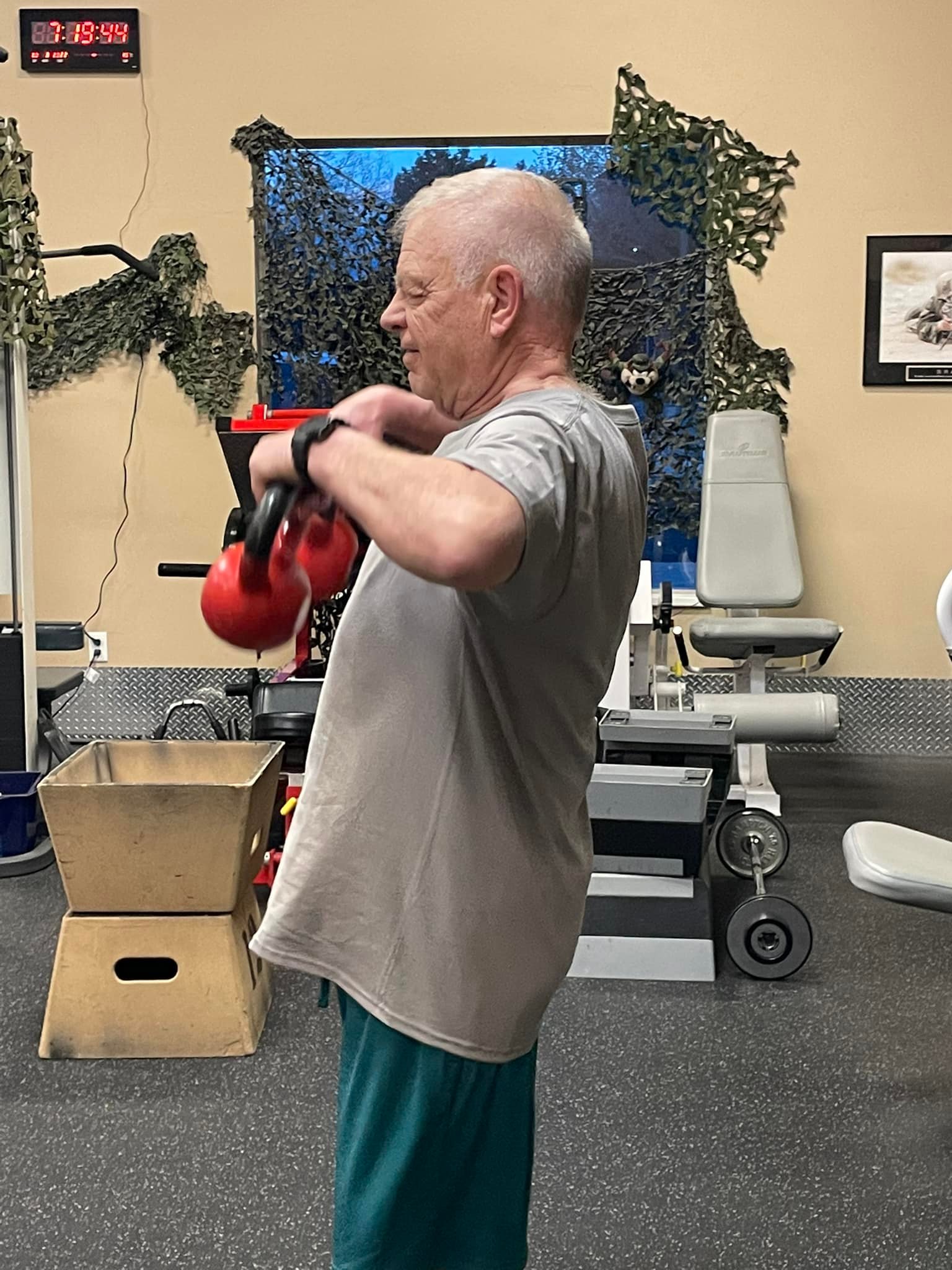 An older man in a gray t-shirt and green shorts lifts a red kettlebell at a gym, surrounded by various exercise equipment and a digital clock on the wall.
