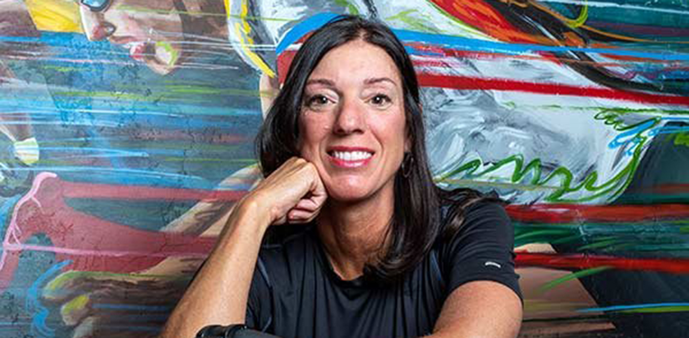 A woman with dark hair wearing a black shirt smiles and rests her chin on her hand in front of a colorful mural featuring an athlete in motion.