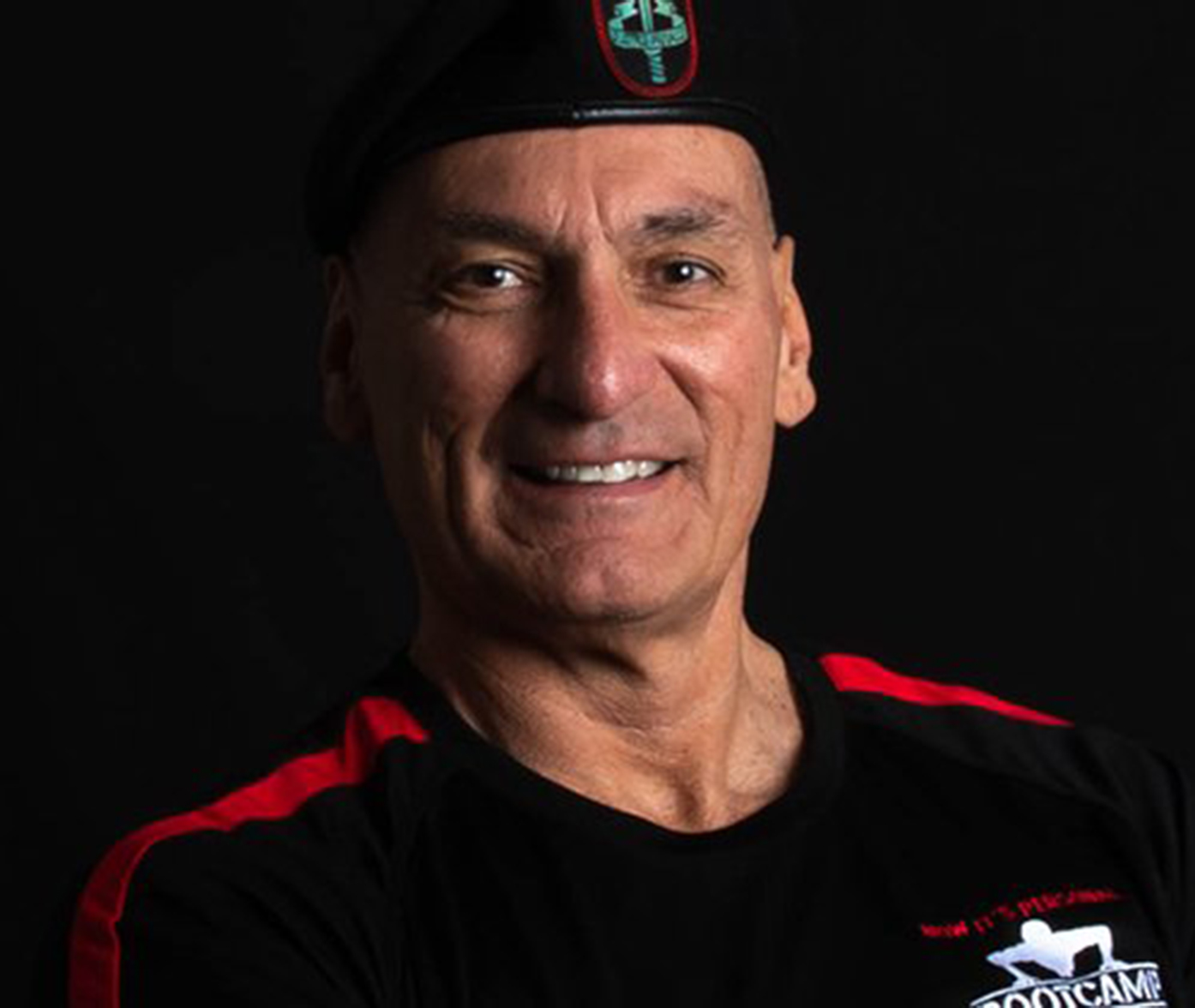 Smiling man in a black beret and black t-shirt with red stripes, posing against a dark background.