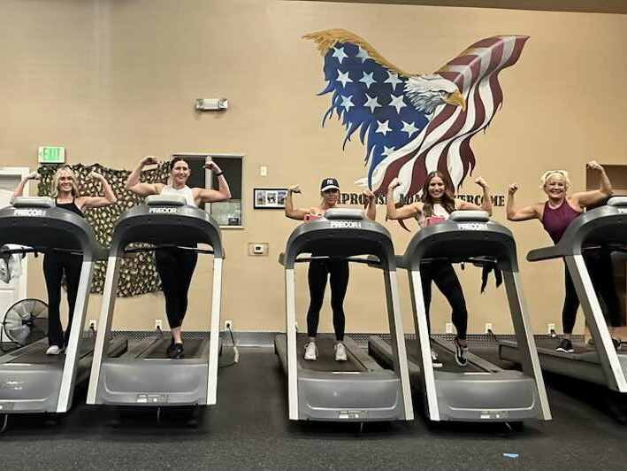 Five women stand on treadmills, flexing their arms and smiling at the camera. Behind them is a mural of a bald eagle and an American flag on the wall. They appear to be in a gym or fitness center.