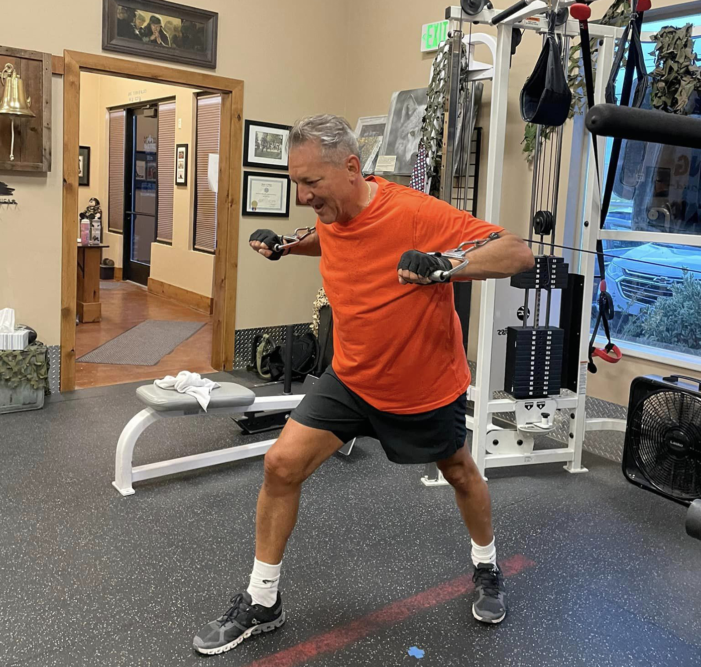 An older man wearing an orange shirt and black shorts exercises with cable machines in a gym, standing in a lunge position and pulling the handles forward with effort. Exercise equipment and a bench are visible in the background.