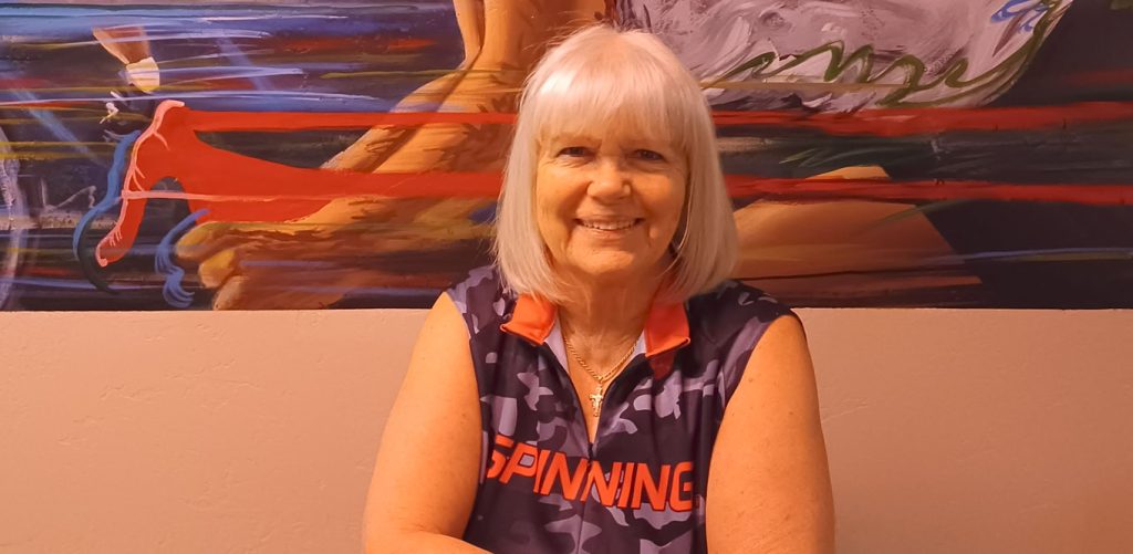 An older woman with gray hair smiles while sitting in front of a colorful mural that features athletic figures. She wears a sleeveless "Spinning" shirt in blue and red tones.